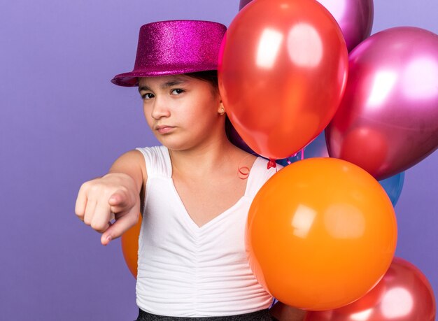 jeune fille caucasienne confiante avec un chapeau de fête violet tenant des ballons à l'hélium et pointant isolé sur un mur violet avec espace de copie