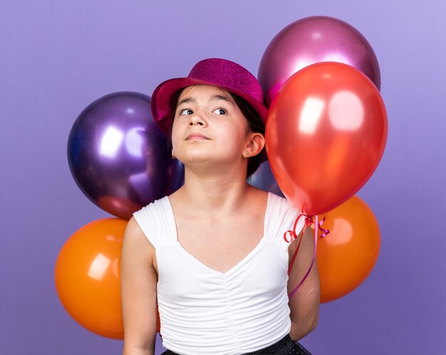 Jeune Fille Caucasienne Confiante Avec Un Chapeau De Fête Violet Tenant Des Ballons à L'hélium Et Regardant Le Côté Isolé Sur Un Mur Violet Avec Espace De Copie