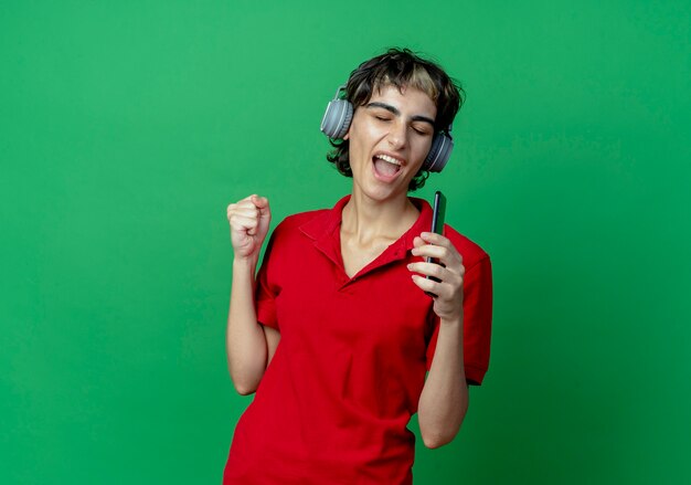Jeune fille caucasienne avec coupe de cheveux de lutin portant des écouteurs écoutant de la musique tenant un téléphone portable prétendant chanter en utilisant le téléphone comme microphone avec le poing fermé et les yeux fermés