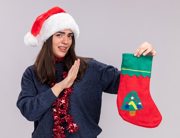 Photo gratuite une jeune fille caucasienne mécontente avec un bonnet de noel et une guirlande autour du cou tient un bas de noël isolé sur un mur blanc avec un espace de copie