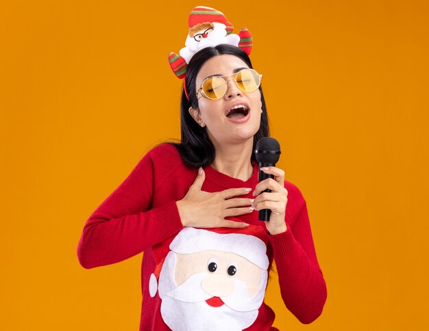 jeune fille caucasienne portant un bandeau de père noël et un pull avec des lunettes tenant un microphone près de la bouche chantant les yeux fermés en gardant la main sur la poitrine isolée sur un mur orange avec un espace de copie