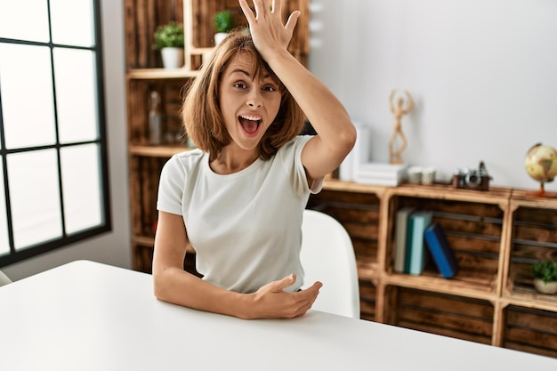 Photo gratuite jeune fille caucasienne portant des vêtements décontractés assise sur la table à la maison surprise avec la main sur la tête pour erreur, rappelez-vous l'erreur oublié le mauvais concept de mémoire