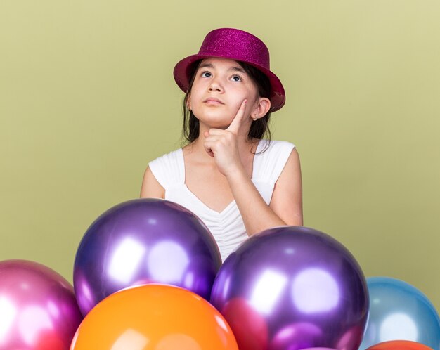 jeune fille caucasienne réfléchie avec un chapeau de fête violet mettant la main sur le menton et levant les yeux debout avec des ballons à l'hélium isolés sur un mur vert olive avec espace de copie