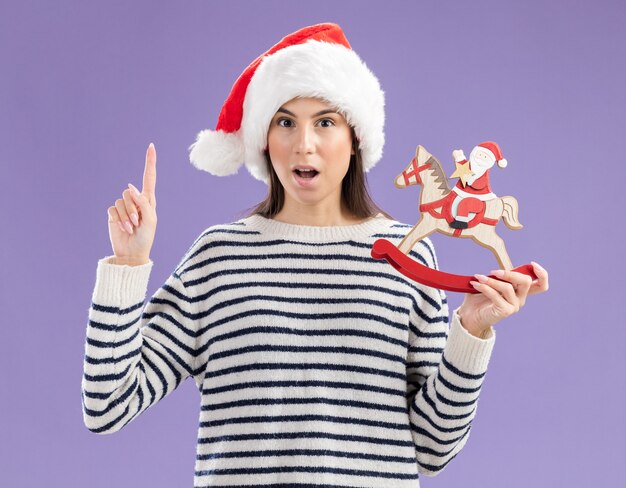 Une jeune fille caucasienne surprise avec un bonnet de noel tient le père Noël sur une décoration de cheval à bascule et le pouce levé isolé sur un mur violet avec espace de copie