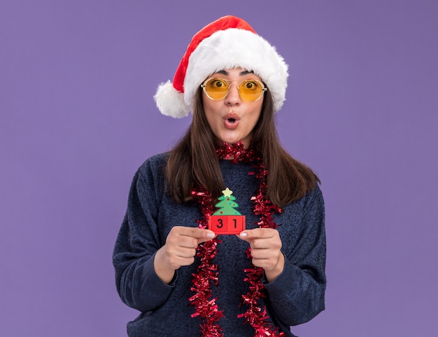 Jeune fille caucasienne surprise en lunettes de soleil avec bonnet de noel et guirlande autour du cou tenant un ornement d'arbre de noël isolé sur un mur violet avec espace pour copie