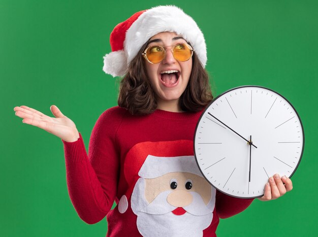 Jeune fille en chandail de Noël portant bonnet de Noel et lunettes regardant heureux et excité tenant horloge murale debout sur le mur vert