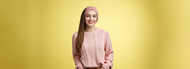 Photo gratuite une jeune fille charmante et sympathique dans un pull tricoté et un bandeau de tête tenant les mains dans les poches souriante