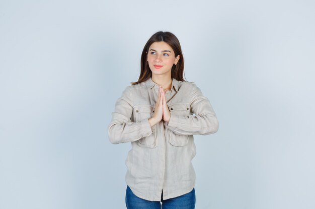 Jeune fille en chemise beige, jeans montrant le geste de namaste et l'air mignon, vue de face.