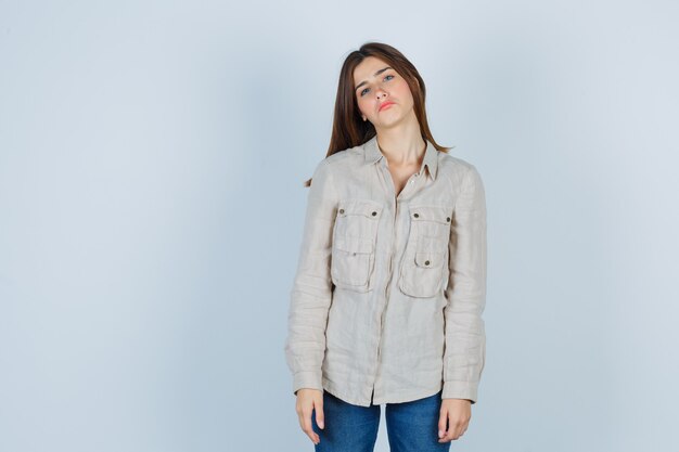 Jeune fille en chemise beige, jeans pliant la tête en posant et l'air mignon, vue de face.