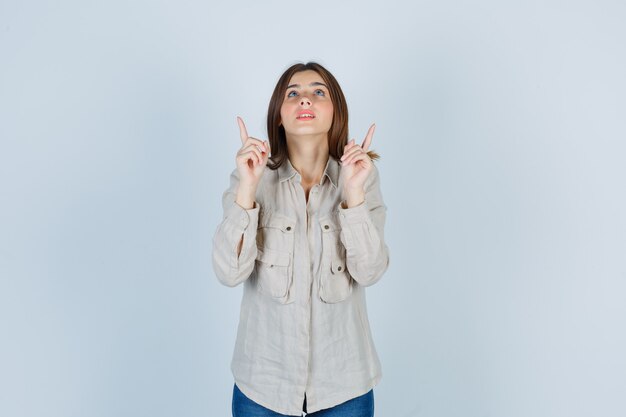 Jeune fille en chemise beige, jeans pointant vers le haut avec l'index et l'air concentré, vue de face.