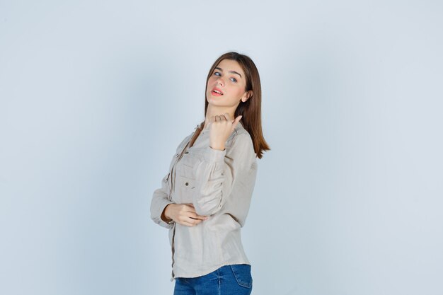 Jeune fille en chemise beige, jeans serrant le poing, regardant par-dessus l'épaule et l'air mignon, vue de face.