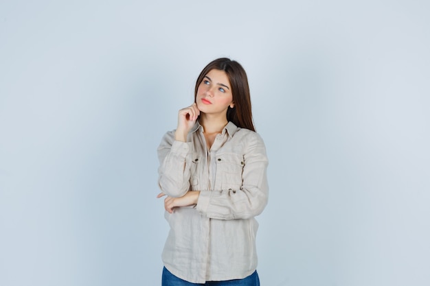 Jeune fille en chemise beige, jeans soutenant le menton à portée de main, pensant à quelque chose et l'air pensif, vue de face.