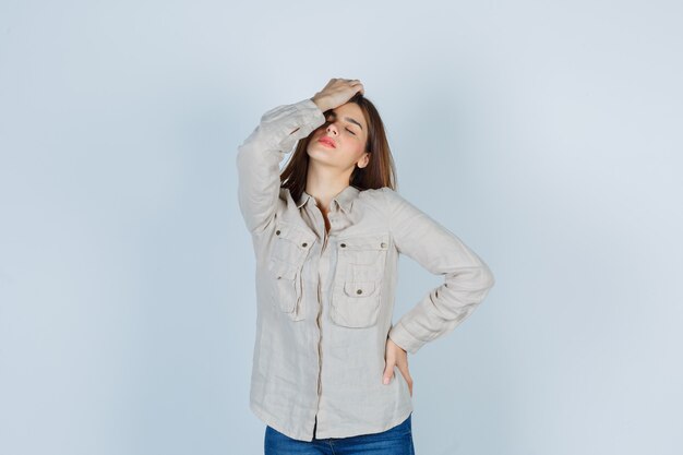 Jeune fille en chemise beige, jeans tenant une main sur la tête, une autre main sur la hanche et l'air harcelé, vue de face.