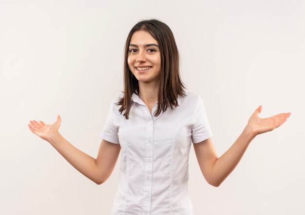 Jeune fille en chemise blanche à l'avant souriant gaiement écartant les bras sur les côtés debout sur un mur blanc