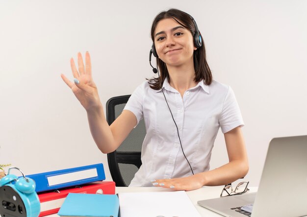 Jeune fille en chemise blanche et casque, montrant le numéro trois avec les doigts souriant assis à la table avec des dossiers et un ordinateur portable sur un mur blanc