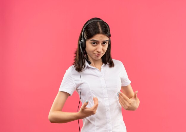 Jeune fille en chemise blanche et casque, pointant avec les bras vers elle-même à la confusion debout sur le mur rose