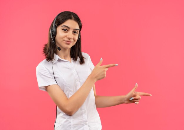 Jeune fille en chemise blanche et casque, pointant avec les doigts sur le côté souriant debout sur le mur rose