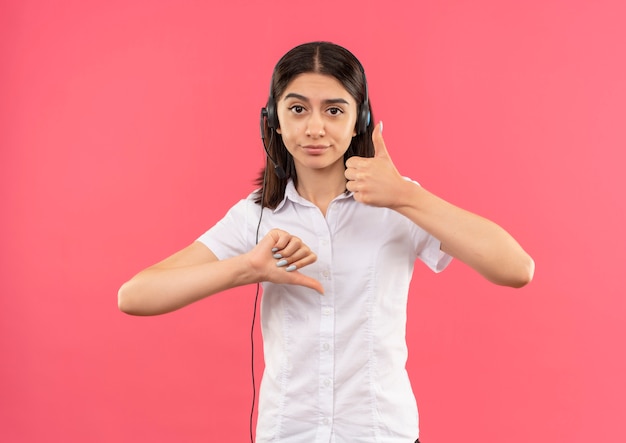 Jeune fille en chemise blanche et casque, regardant vers l'avant montrant les pouces de haut en bas debout sur un mur rose