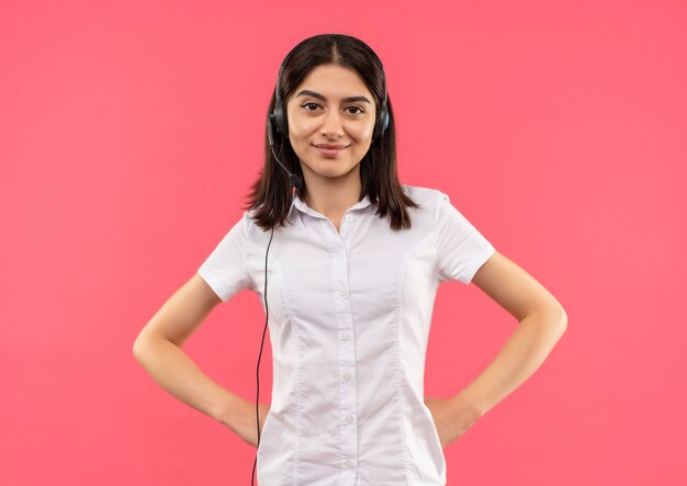 Jeune fille en chemise blanche et casque, regardant vers l'avant en souriant avec une expression confiante debout sur un mur rose