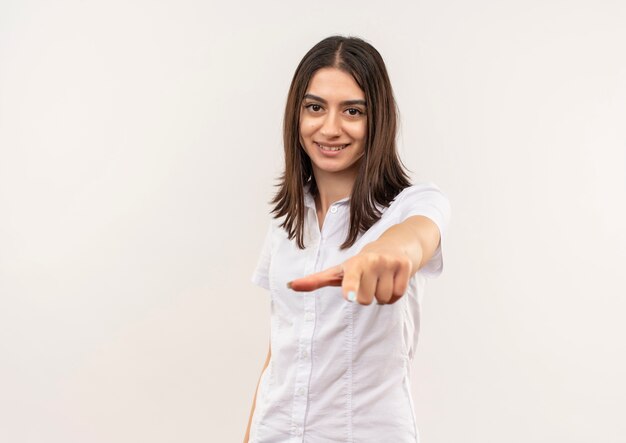 Jeune fille en chemise blanche pointant avec le doigt vers l'avant souriant confiant debout sur un mur blanc