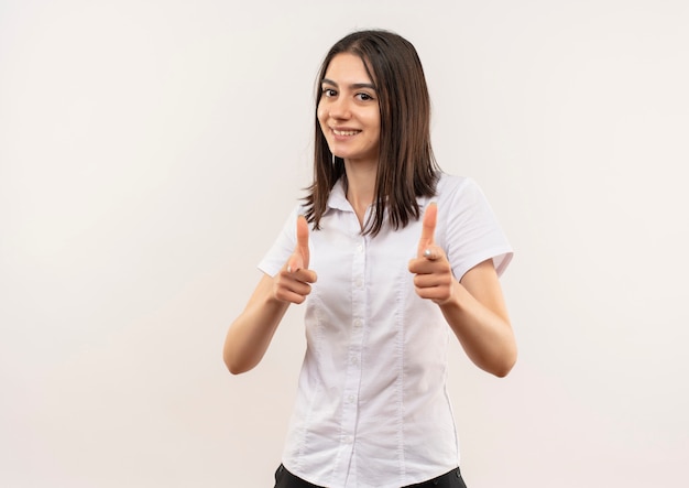 Jeune fille en chemise blanche pointant avec l'index vers l'avant souriant joyeusement debout sur un mur blanc