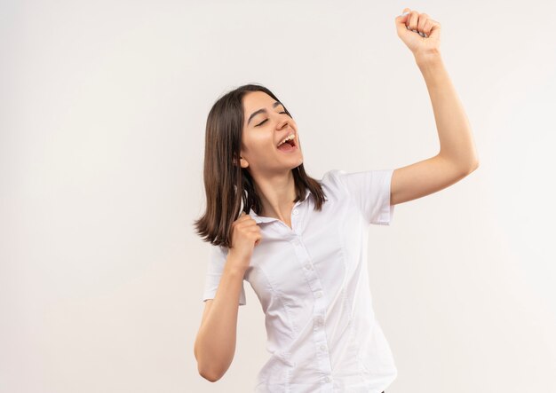 Jeune fille en chemise blanche serrant le poing se réjouissant de son succès heureux et excité debout sur un mur blanc