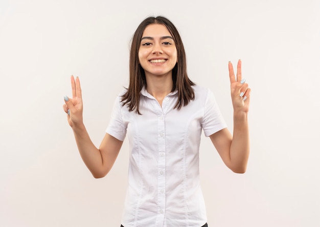 Jeune fille en chemise blanche souriant joyeusement montrant la victoire chanter avec les deux mains debout sur un mur blanc