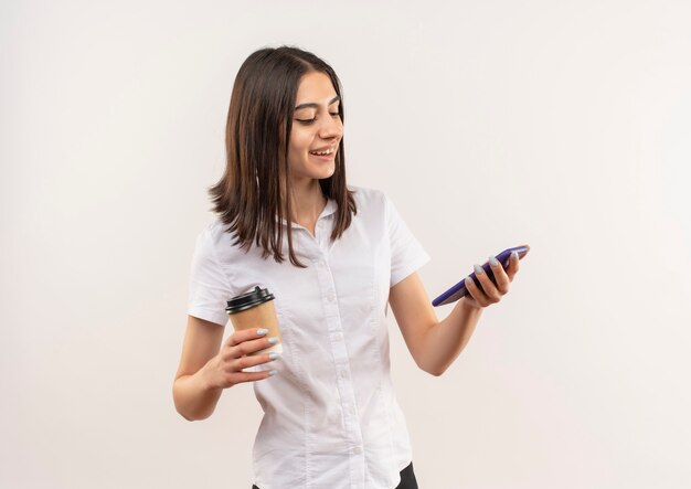 Jeune fille en chemise blanche tenant une tasse de café en regardant l'écran de son mobile souriant joyeusement debout sur un mur blanc