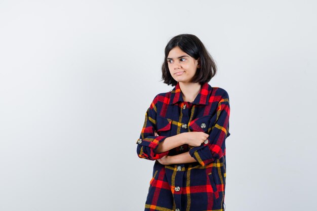 Jeune fille en chemise à carreaux debout les bras croisés, regardant loin et l'air heureux, vue de face.