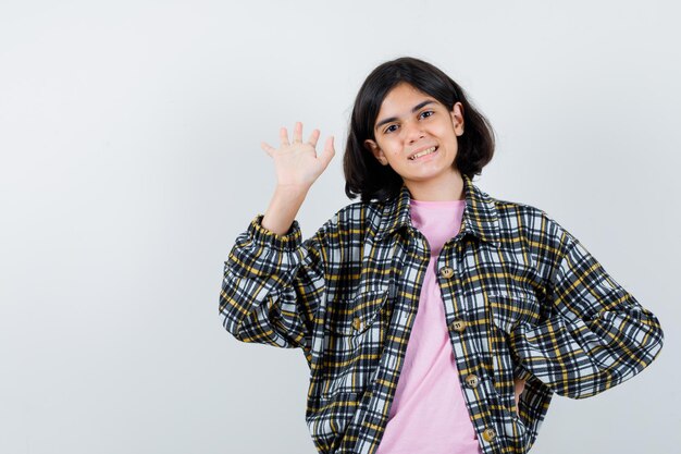 Jeune fille en chemise à carreaux et t-shirt rose étirant la main en saluant quelqu'un tout en tenant la main sur la taille et en ayant l'air mignon