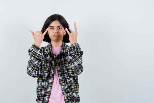 Photo gratuite jeune fille en chemise à carreaux et t-shirt rose montrant le geste rock n roll et le petit doigt et l'air sérieux, vue de face.