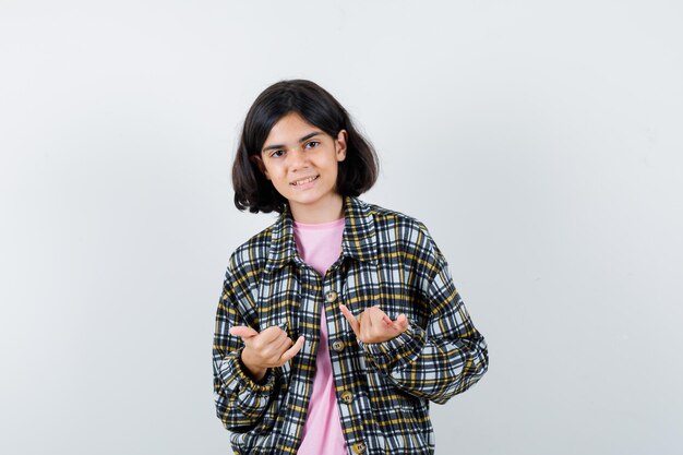 Jeune fille en chemise à carreaux et t-shirt rose montrant le petit doigt et le geste rock n roll et l'air mignon, vue de face.