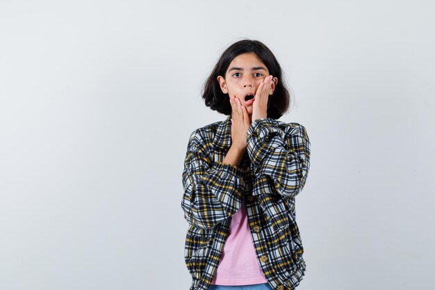 Jeune fille en chemise à carreaux et t-shirt rose tenant la main près de la bouche et l'air surpris, vue de face.