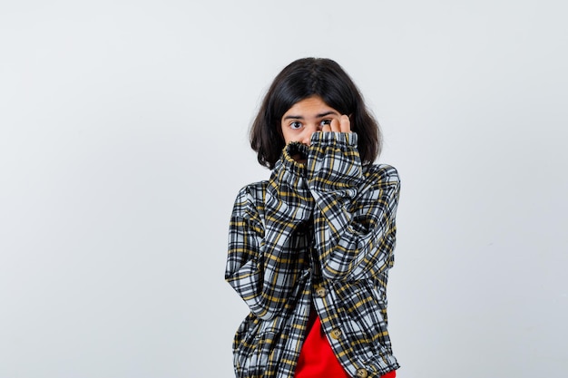 Jeune fille en chemise à carreaux et t-shirt rouge couvrant le visage avec les mains et ayant l'air effrayé, vue de face.