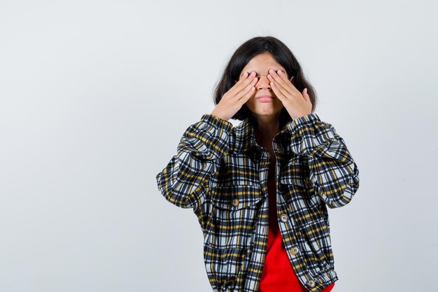 Jeune fille en chemise à carreaux et t-shirt rouge couvrant les yeux avec les mains et l'air sérieux, vue de face.