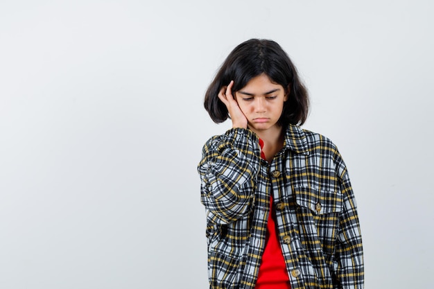 Jeune fille en chemise à carreaux et t-shirt rouge, la joue appuyée sur la paume et l'air sérieux, vue de face.