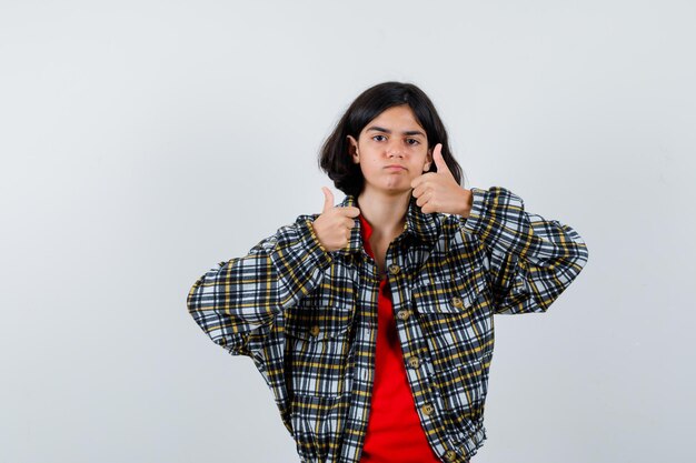 Jeune fille en chemise à carreaux et t-shirt rouge montrant les pouces vers le haut avec les deux mains et l'air mignon, vue de face.