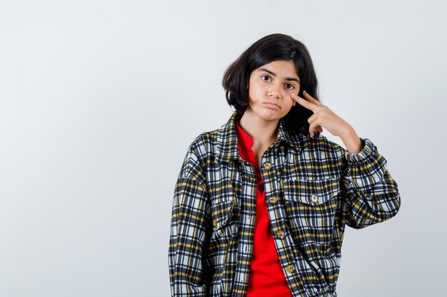 Jeune fille en chemise à carreaux et t-shirt rouge montrant le signe v et l'air sérieux, vue de face.