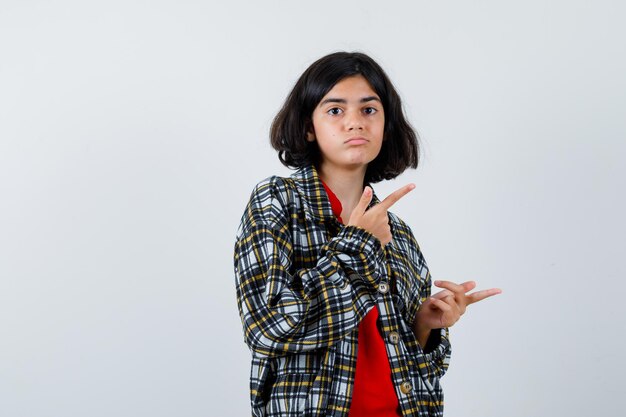 Jeune fille en chemise à carreaux et t-shirt rouge pointant vers la droite avec l'index et l'air sérieux, vue de face.