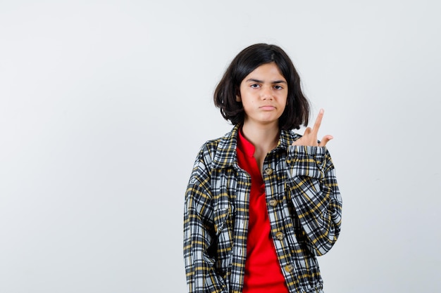 Jeune fille en chemise à carreaux et t-shirt rouge pointant vers le haut et l'air sérieux, vue de face.