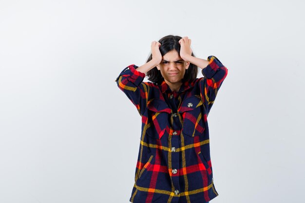 Jeune fille en chemise à carreaux tenant les mains sur la tête et l'air anxieux, vue de face.