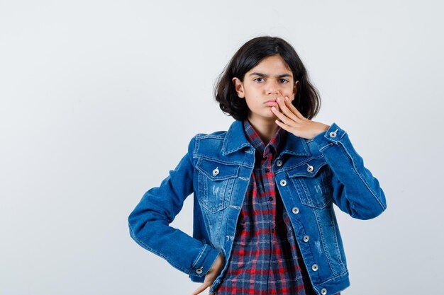 Jeune fille en chemise à carreaux et veste en jean couvrant la bouche avec la main tout en tenant la main sur la taille et l'air pensif, vue de face.