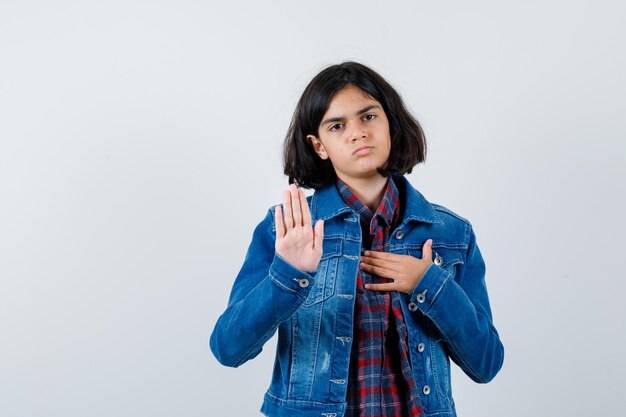 Jeune fille en chemise à carreaux et veste en jean montrant un panneau d'arrêt tout en posant la main sur la poitrine et l'air sérieux