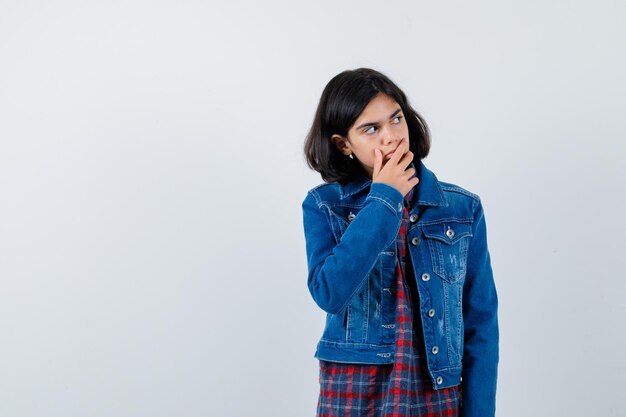 Jeune fille en chemise à carreaux et veste en jean regardant loin et posant à la caméra et regardant pensive, vue de face.