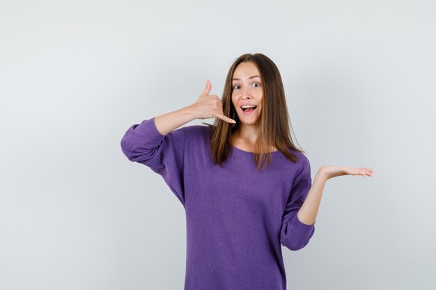 Jeune fille en chemise violette montrant le signe du téléphone avec la paume de côté et à la vue utile, de face.