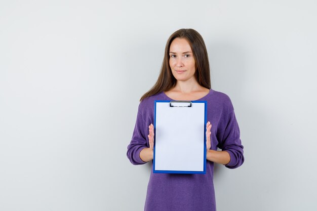 Jeune fille en chemise violette tenant le presse-papiers et souriant, vue de face.