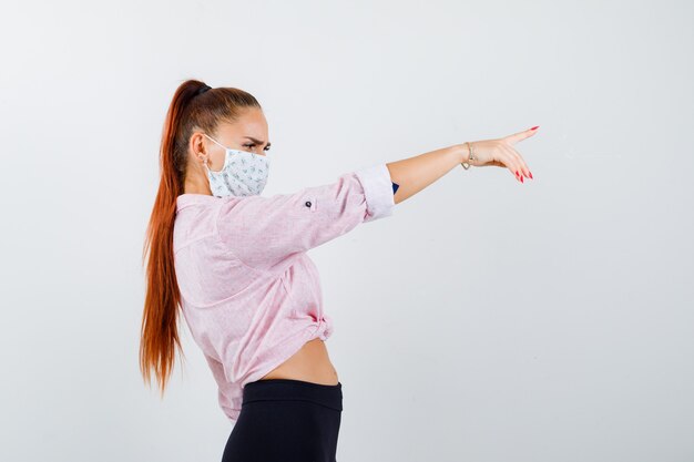 Jeune fille en chemisier rose, pantalon noir, masque pointant vers la droite avec l'index et regardant focalisé, vue de face.