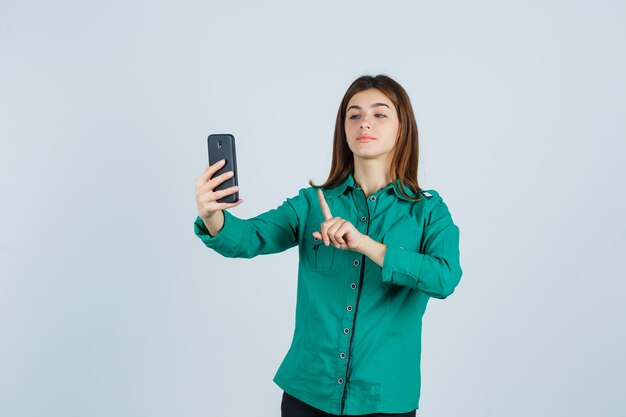 Jeune fille en chemisier vert, pantalon noir montrant tenir sur un geste minute tout en faisant un appel vidéo et à la jolie vue de face.