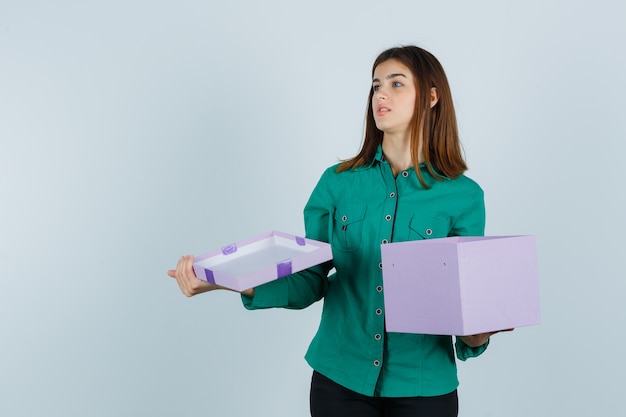 Jeune fille en chemisier vert, pantalon noir ouvrant la boîte-cadeau, regardant ailleurs et regardant focalisé, vue de face