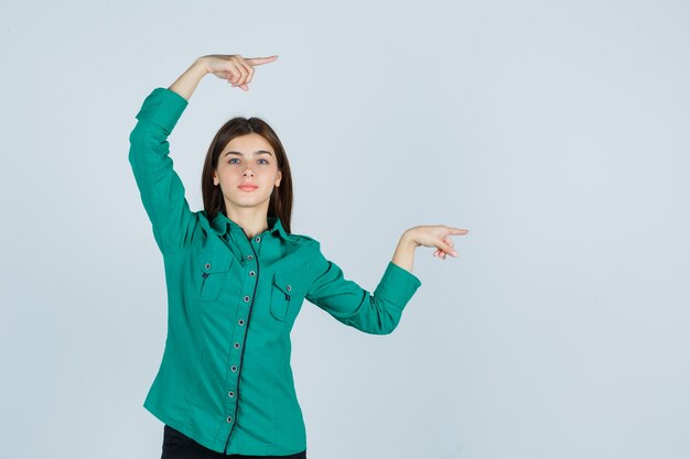 Jeune fille en chemisier vert, pantalon noir pointant vers la droite avec l'index et l'air heureux, vue de face.